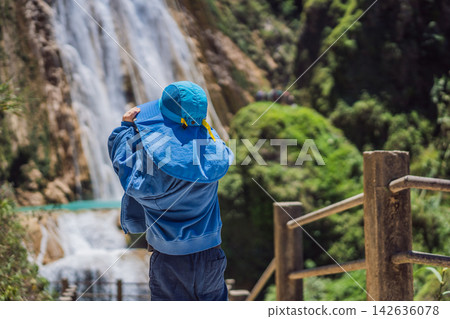 Little boy tourist looking at El Chiflon waterfall and turquoise water in Chiapas Mexico with wooden railings tropical forest clear blue water nature travel and walking. Little boy tourist looking at El Chiflon waterfall and turquoise water in Chiapas Mexico with wooden railings tropical forest clear blue water nature travel and walking. 142636078