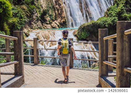 Boy tourist looking at El Chiflon waterfall and turquoise water in Chiapas Mexico with wooden railings tropical forest clear blue water nature travel and rear view. Boy tourist looking at El Chiflon waterfall and turquoise water in Chiapas Mexico with wooden railings tropical forest clear blue water nature travel and rear view. 142636081