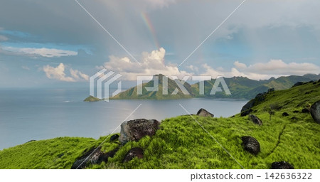 Lush green hills and volcanic mountains in the Fiji islands rising from calm turquoise ocean waters under a cloudy sky with a vibrant rainbow arching over the distant peaks Lush green hills and volcanic mountains in the Fiji islands rising from calm turquoise ocean waters under a cloudy sky with a vibrant rainbow arching over the distant peaks 142636322