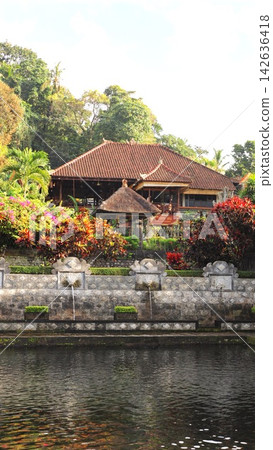 Tropical resort hotel rises behind decorative stone fountains and dense greenery, in Balinese style. Resort hotel overlooks still water, with red roof tiles, garden plants, and tranquil atmosphere. 142636418