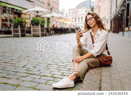 Tourist wearing glasses, holding phone in hands walk around city at sunset. Travel, blogging concept Tourist wearing glasses, holding phone in hands walk around city at sunset. Travel, blogging concept 142636456