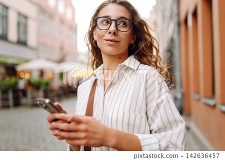 Tourist wearing glasses, holding phone in hands walk around city at sunset. Travel, blogging concept Tourist wearing glasses, holding phone in hands walk around city at sunset. Travel, blogging concept 142636457