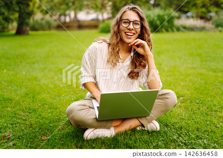Young woman in glasses and with laptop working at sunset in park on green meadow. Lifestyle concept. Young woman in glasses and with laptop working at sunset in park on green meadow. Lifestyle concept. 142636458
