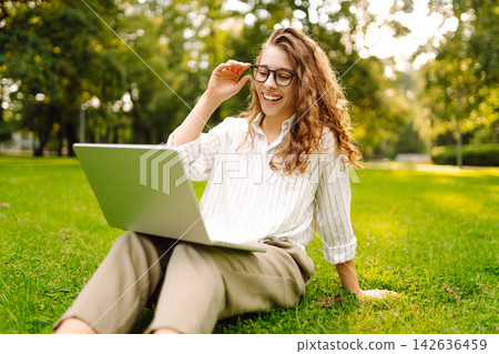 Young woman in glasses and with laptop working at sunset in park on green meadow. Lifestyle concept. Young woman in glasses and with laptop working at sunset in park on green meadow. Lifestyle concept. 142636459