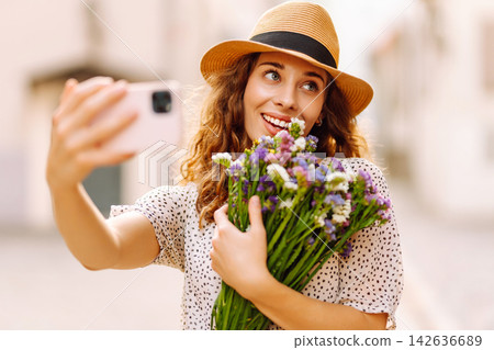 Woman with bouquet of wild flowers taking selfie on phone against city street. Blogging concept. Woman with bouquet of wild flowers taking selfie on phone against city street. Blogging concept. 142636689