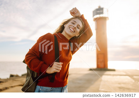 Young woman enjoying a breezy day near a vibrant red lighthouse. Autumn sunset 142636917