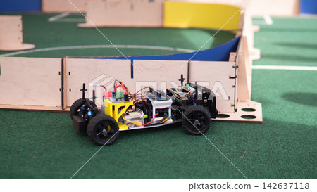 Compact four wheeled robot car with visible wiring, sensors and circuit boards moves on green turf inside wooden course, showing indoor robotics testing, engineering prototype, and autonomous Compact four wheeled robot car with visible wiring, sensors and circuit boards moves on green turf inside wooden course, showing indoor robotics testing, engineering prototype, and autonomous 142637118