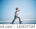 A barefoot boy playing by throwing seashells at the water's edge on a summer beach. 142637184