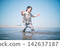 A low-angle shot of a boy smiling in the shallow waters of a summer beach, holding a seashell. 142637187