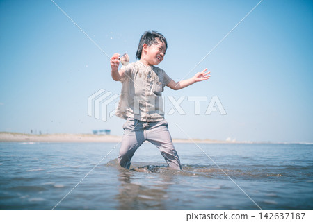 A low-angle shot of a boy smiling in the shallow waters of a summer beach, holding a seashell. A low-angle shot of a boy smiling in the shallow waters of a summer beach, holding a seashell. 142637187