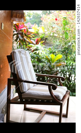 Empty wooden lounge chair with striped cushions sits beside vibrant tropical greenery on tiled patio. Patio armchair provides comfortable outdoor seating with broad arms and sunlit plants. Empty wooden lounge chair with striped cushions sits beside vibrant tropical greenery on tiled patio. Patio armchair provides comfortable outdoor seating with broad arms and sunlit plants. 142637414