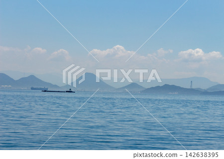 A distant view of the Seto Inland Sea, showing a cargo ship and a barge sailing on calm waters, with the mountain range on the opposite shore (Kagawa Prefecture) in the background. A distant view of the Seto Inland Sea, showing a cargo ship and a barge sailing on calm waters, with the mountain range on the opposite shore (Kagawa Prefecture) in the background. 142638395