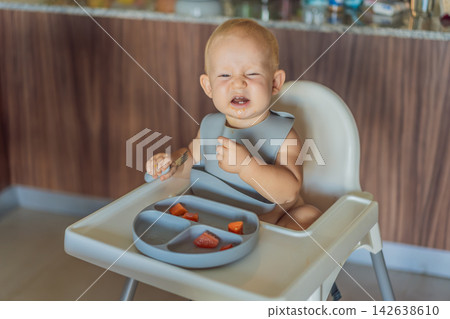 Infant practices self feeding with vegetables in a kitchen, showing baby led weaning, early nutrition and family care concept Infant practices self feeding with vegetables in a kitchen, showing baby led weaning, early nutrition and family care concept 142638610