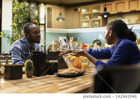 African american man working too hard and woman asking for a break, showing him the timeout sign and closing his laptop. Workaholic boyfriend ignoring his girlfriend during breakfast. African american man working too hard and woman asking for a break, showing him the timeout sign and closing his laptop. Workaholic boyfriend ignoring his girlfriend during breakfast. 142642048