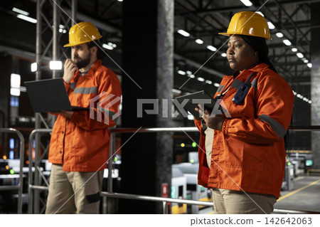 Black woman staff, checking factory floor, heavy machinery, industrial space filled with steel work and mechanical systems. Personnel overseeing heavy industry engineering activity. Black woman staff, checking factory floor, heavy machinery, industrial space filled with steel work and mechanical systems. Personnel overseeing heavy industry engineering activity. 142642063