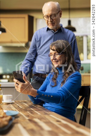 Senior woman and her husband laughing and checking smartphone at the breakfast table to enjoy a cozy meal with toast and coffee. Chatting and reflecting warm retirement lifestyle. Senior woman and her husband laughing and checking smartphone at the breakfast table to enjoy a cozy meal with toast and coffee. Chatting and reflecting warm retirement lifestyle. 142642293