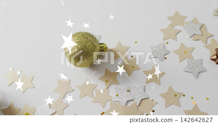 Resting gold glitter bauble on white flatlay, surrounding beige and silver star confetti, metal bow 142642627