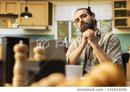 Man cracking knuckles, nervous habit at home, repetitive involuntary popping sound, focused on tasks, Using laptop in the kitchen, teleworking and multitasking at the wooden table. Man cracking knuckles, nervous habit at home, repetitive involuntary popping sound, focused on tasks, Using laptop in the kitchen, teleworking and multitasking at the wooden table. 142643046
