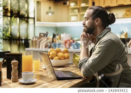 Adult male meditating and contemplating for mental wellness, working remotely on laptop in the kitchen. Digital nomad relaxed and zen during meditation break, eyes closed. Adult male meditating and contemplating for mental wellness, working remotely on laptop in the kitchen. Digital nomad relaxed and zen during meditation break, eyes closed. 142643411