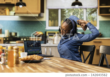 Video editor woman stretching her muscles after assembling montage on laptop, video production and timeline editing. Digital nomad videographer with cramps and muscle stiffness at table. Video editor woman stretching her muscles after assembling montage on laptop, video production and timeline editing. Digital nomad videographer with cramps and muscle stiffness at table. 142643562