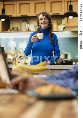 Portrait of mature housewife standing in her apartment kitchen, smiling with calm enjoyment. Elder adult embracing retired life with natural household chores, creating a positive domestic living. 142643855