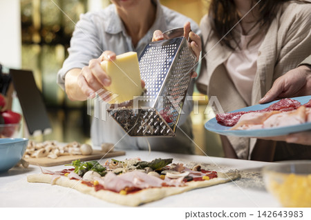 Close up of mother grating cheese on a grater for homemade pizza, cooking with pre-teen daughter and husband. Adding grated mozzarella sprinkles on top of dough and ingredients. Close up of mother grating cheese on a grater for homemade pizza, cooking with pre-teen daughter and husband. Adding grated mozzarella sprinkles on top of dough and ingredients. 142643983
