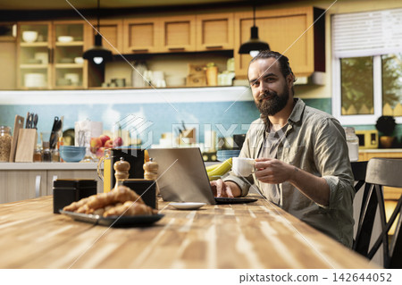 Portrait of male digital nomad using his laptop at the kitchen table, enjoying relaxed freelancer lifestyle with flexible work. Adult man telecommuting indoors, setup for productivity. Portrait of male digital nomad using his laptop at the kitchen table, enjoying relaxed freelancer lifestyle with flexible work. Adult man telecommuting indoors, setup for productivity. 142644052