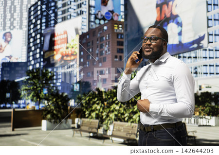 African american adult man on a city street answering a phone call, captures independence and modernity surrounded by sunlight and the scenery of a busy metropolitan environment. African american adult man on a city street answering a phone call, captures independence and modernity surrounded by sunlight and the scenery of a busy metropolitan environment. 142644203