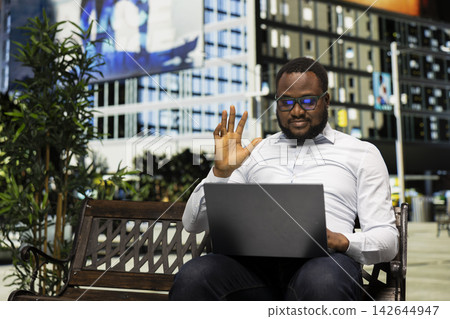 Confident African American man waving during video call on laptop, sitting on a bench enjoying a casual online meeting in a vibrant downtown setting. Urban architecture and active city life. Confident African American man waving during video call on laptop, sitting on a bench enjoying a casual online meeting in a vibrant downtown setting. Urban architecture and active city life. 142644947