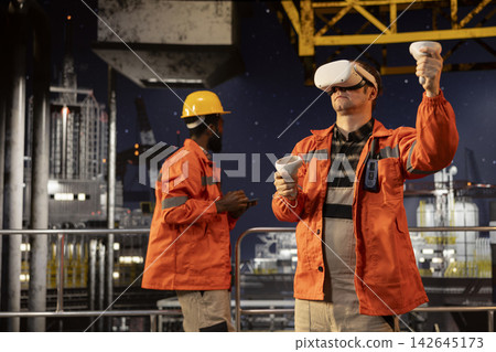 Industrial male employee on offshore platform wears virtual reality gear to oversee oil and gas drilling simulation, using augmented technology for advanced modern rig maintenance. Industrial male employee on offshore platform wears virtual reality gear to oversee oil and gas drilling simulation, using augmented technology for advanced modern rig maintenance. 142645173