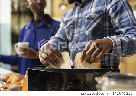 Boyfriend and girlfriend preparing bread in a toaster together for breakfast table, enjoying toast, homemade pastry and morning coffee. Couple showing love and fun in a warm inviting kitchen. Boyfriend and girlfriend preparing bread in a toaster together for breakfast table, enjoying toast, homemade pastry and morning coffee. Couple showing love and fun in a warm inviting kitchen. 142645235