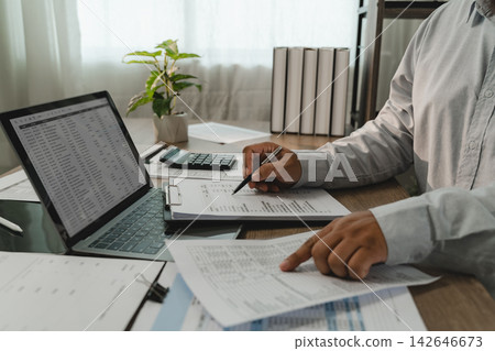 Businessman reviewing financial report beside laptop spreadsheet, comparing printed figures with accounting data for financial analysis and reporting workflow. Businessman reviewing financial report beside laptop spreadsheet, comparing printed figures with accounting data for financial analysis and reporting workflow. 142646673