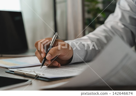 Close-up of a hand holding a pen over printed agreement documents on a clipboard beside a laptop, showing contract terms review and approval preparation in an office. Close-up of a hand holding a pen over printed agreement documents on a clipboard beside a laptop, showing contract terms review and approval preparation in an office. 142646674