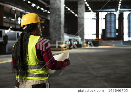 Black woman worker analyzes blueprints and construction drawings inside a spacious industrial warehouse. Production hall showcases metalwork, steel beams and advanced machinery for production. Black woman worker analyzes blueprints and construction drawings inside a spacious industrial warehouse. Production hall showcases metalwork, steel beams and advanced machinery for production. 142646705