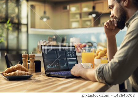 Remote administrator, laptop displaying analytics, charts and research data, working indoors at kitchen table. Lifestyle of telecommuting, task management and productivity, independent work. Remote administrator, laptop displaying analytics, charts and research data, working indoors at kitchen table. Lifestyle of telecommuting, task management and productivity, independent work. 142646708