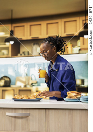 African american girl standing in the kitchen with a glass of juice, looking at delicious croissants on a plate. Morning routine expresses calmness and happiness in comfortable atmosphere. African american girl standing in the kitchen with a glass of juice, looking at delicious croissants on a plate. Morning routine expresses calmness and happiness in comfortable atmosphere. 142646714