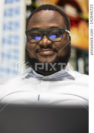 Portrait of black man sitting on a bench with a laptop navigating online websites, enjoying sunlight in a small downtown park. Modern urban architecture with busy city street vibes. Portrait of black man sitting on a bench with a laptop navigating online websites, enjoying sunlight in a small downtown park. Modern urban architecture with busy city street vibes. 142646723