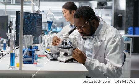 Researcher operating compound microscope, doing sample observation in controlled laboratory environment. African american man studies tissue under optical device in biotechnology lab, camera B Researcher operating compound microscope, doing sample observation in controlled laboratory environment. African american man studies tissue under optical device in biotechnology lab, camera B 142646765