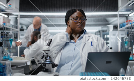 Tired clinical laboratory technologist feeling neck ache while working on laptop, studying genomics. African american woman relaxes strained muscles while studying DNA strand mapping, camera A Tired clinical laboratory technologist feeling neck ache while working on laptop, studying genomics. African american woman relaxes strained muscles while studying DNA strand mapping, camera A 142646788