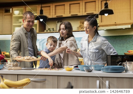 Family prepares homemade meal with excited kids arranging fresh ingredients on the pizza. Teamwork in the kitchen creates a warm lifestyle moment full of laughter and delicious food. Family prepares homemade meal with excited kids arranging fresh ingredients on the pizza. Teamwork in the kitchen creates a warm lifestyle moment full of laughter and delicious food. 142646789
