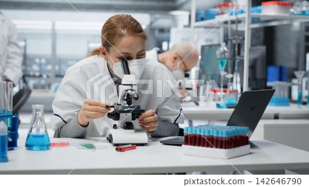 Technician wearing surgical mask examining pathology slides under microscope in research center. Researcher using protective medical gear driving medical innovation, doing cellular analysis, camera B Technician wearing surgical mask examining pathology slides under microscope in research center. Researcher using protective medical gear driving medical innovation, doing cellular analysis, camera B 142646790