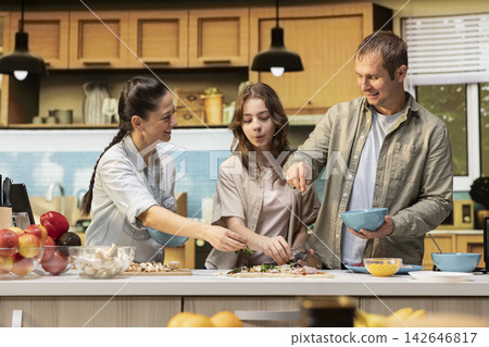 Family making homemade pizza recipe together by adding ingredients in the kitchen, enjoying laughter during fun weekend activity. Teamwork and bonding time with pre-teen daughter. Family making homemade pizza recipe together by adding ingredients in the kitchen, enjoying laughter during fun weekend activity. Teamwork and bonding time with pre-teen daughter. 142646817