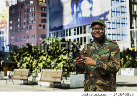 Portrait of military personnel using his phone for call to give patrol orders in metropolis, walking along downtown street wearing tactical camouflage uniform. Honor, courage and readiness. Portrait of military personnel using his phone for call to give patrol orders in metropolis, walking along downtown street wearing tactical camouflage uniform. Honor, courage and readiness. 142646856
