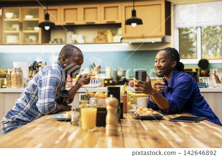 Black people checking something on smartphone enjoying breakfast at home, sitting at the kitchen table. Drinking fresh coffee together and chatting over croissants for morning routine. 142646892
