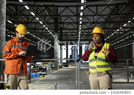 Black woman professional coordinating industrial operations with walkie-talkie, ensuring two-way radio communication between colleagues for efficient heavy industry manufacturing activity. 142646903