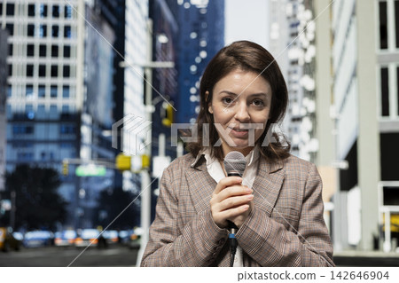 Portrait of cheerful field reporter uses mic and speaks to camera on city street, presents information. Smiling journalist reports live news update with microphone from metropolitan boulevard, closeup 142646904