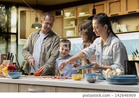 Happy young parents teaching kids how to cook a homemade pizza, explaining steps and ingredients list before cooking together. Family for four bonding during culinary activity. Happy young parents teaching kids how to cook a homemade pizza, explaining steps and ingredients list before cooking together. Family for four bonding during culinary activity. 142646909