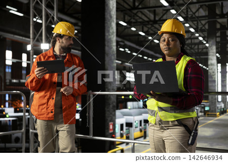 Engineer in hard hat reviewing production line near heavy equipment and metallic beams. Industrial warehouse environment reflects engineering and steel work in machinery hall. Engineer in hard hat reviewing production line near heavy equipment and metallic beams. Industrial warehouse environment reflects engineering and steel work in machinery hall. 142646934