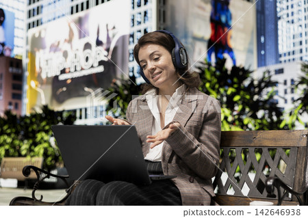 Smiling businesswoman in videocall communicating with coworkers from city outdoors. Happy remote worker using laptop and audio headphones to participates in virtual meeting with colleagues Smiling businesswoman in videocall communicating with coworkers from city outdoors. Happy remote worker using laptop and audio headphones to participates in virtual meeting with colleagues 142646938