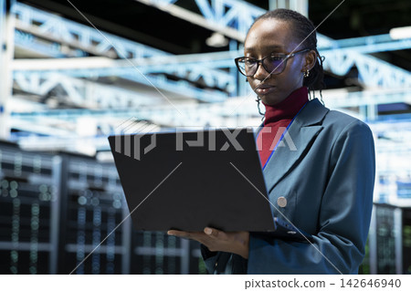 Data center female technician using laptop software to evaluate infrastructure diagnostic reports. African american woman in server hub using notebook application to automate supercomputers processing Data center female technician using laptop software to evaluate infrastructure diagnostic reports. African american woman in server hub using notebook application to automate supercomputers processing 142646940